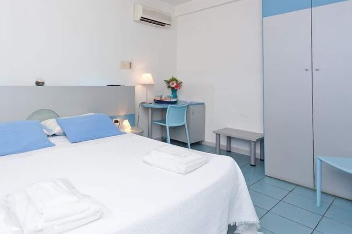 A hotel room with a bed, a desk, a chair, a small table, and a wardrobe.