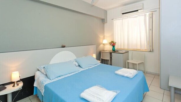 Une chambre d’hôtel comprenant un lit, un bureau, une chaise, une fenêtre avec des rideaux et une table de chevet avec une lampe.