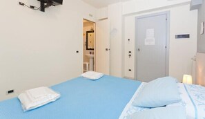 A hotel room with a single bed, a blue mattress, white bedding, a white towel on the bed, a nightstand with a lamp, and a door leading to a bathroom. - Rainbow (Palermo)