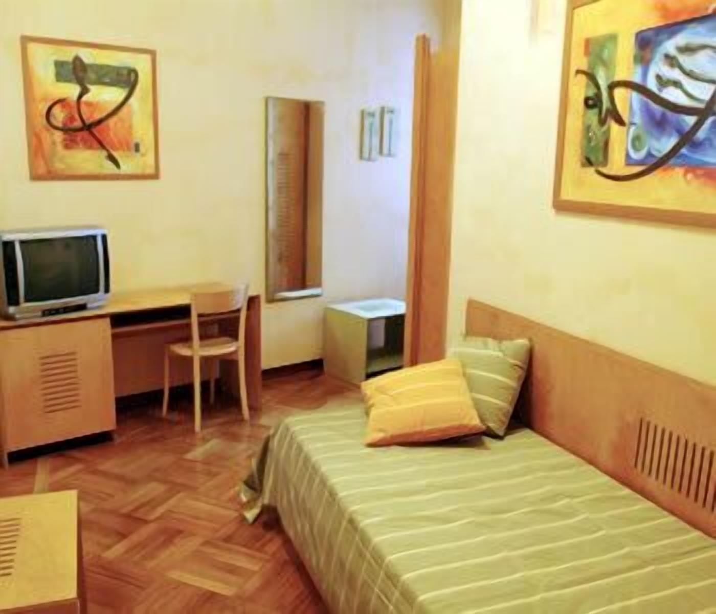 A hotel room with a bed, a desk with a computer, a chair, a small table, and a mirror.