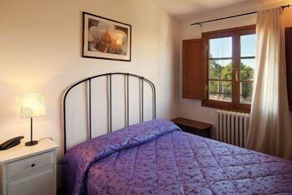 A bedroom with a bed, a nightstand, a lamp, a window with curtains, and a painting on the wall.
