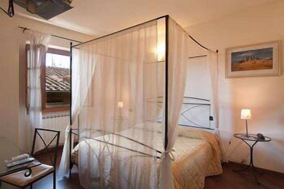 A room with a bed under a mosquito net, a small table with a lamp, and a painting on the wall.