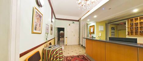 A hotel reception area with a reception desk, chairs, a patterned rug, and framed artwork on the walls.