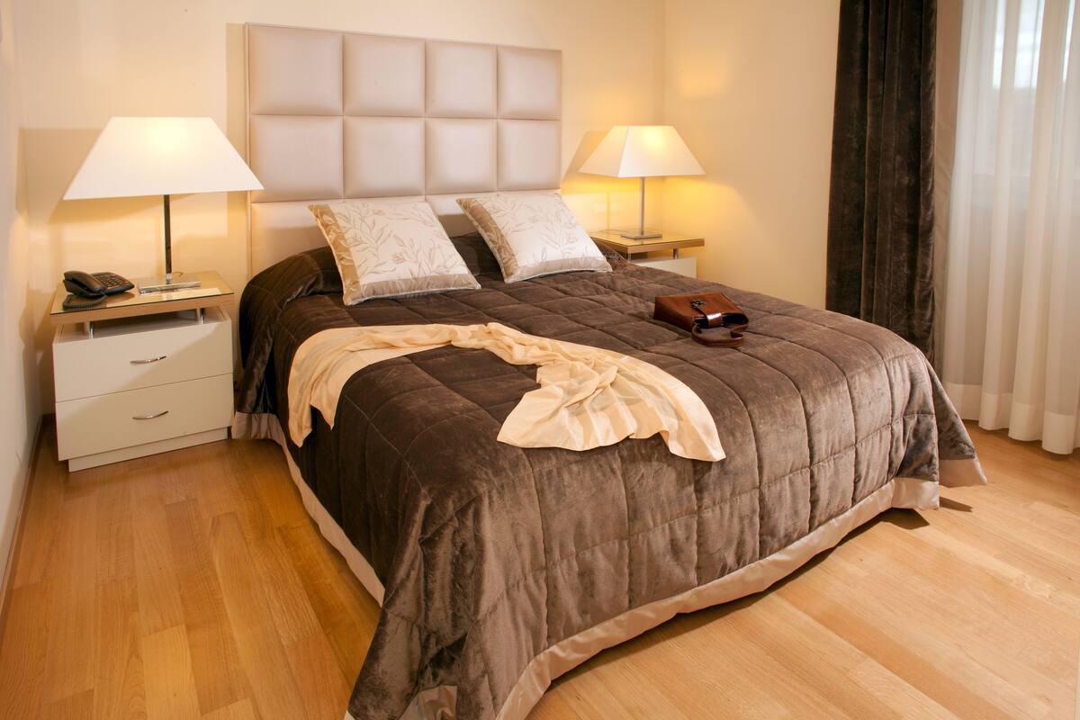 A neatly made bed with a brown duvet, two bedside lamps, a bedside phone, and a small bedside table with a lamp.