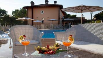 A poolside setting with wicker lounge chairs, a glass table with fruit and drinks, and umbrellas.