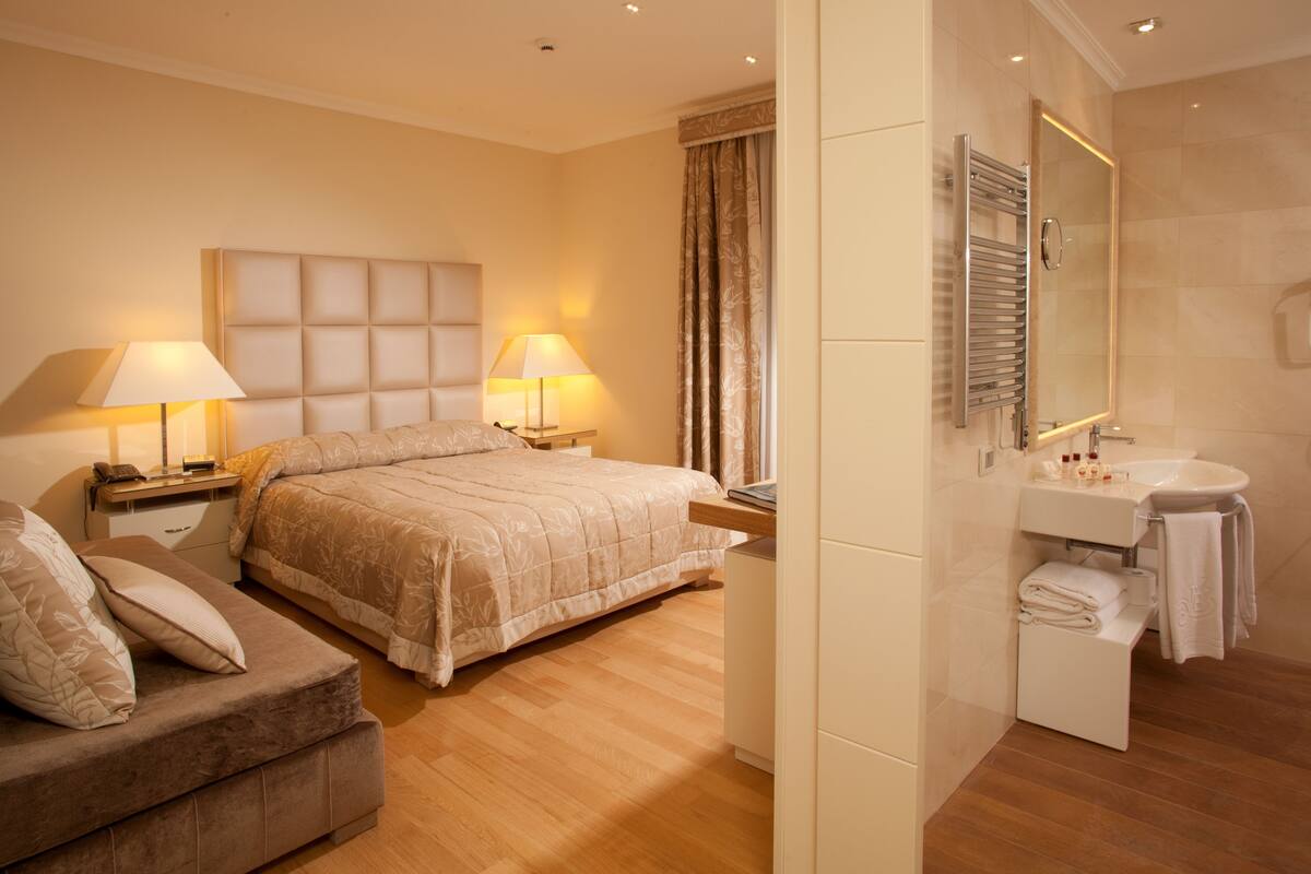 A hotel room with a bed, bedside tables, a sofa, a bathroom with a sink and mirror.