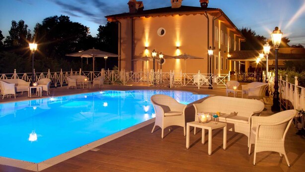 Une piscine entourée de chaises longues et de parasols, le tout délimité par une clôture blanche, et un bâtiment doté de nombreuses fenêtres et de luminaires.