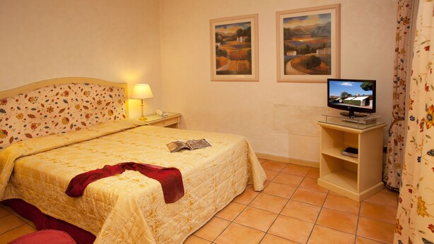 A neatly made bed with a floral headboard, a bedside lamp, a television on a stand, and a small shelf with a book. - Piccolo Borgo (Rome)