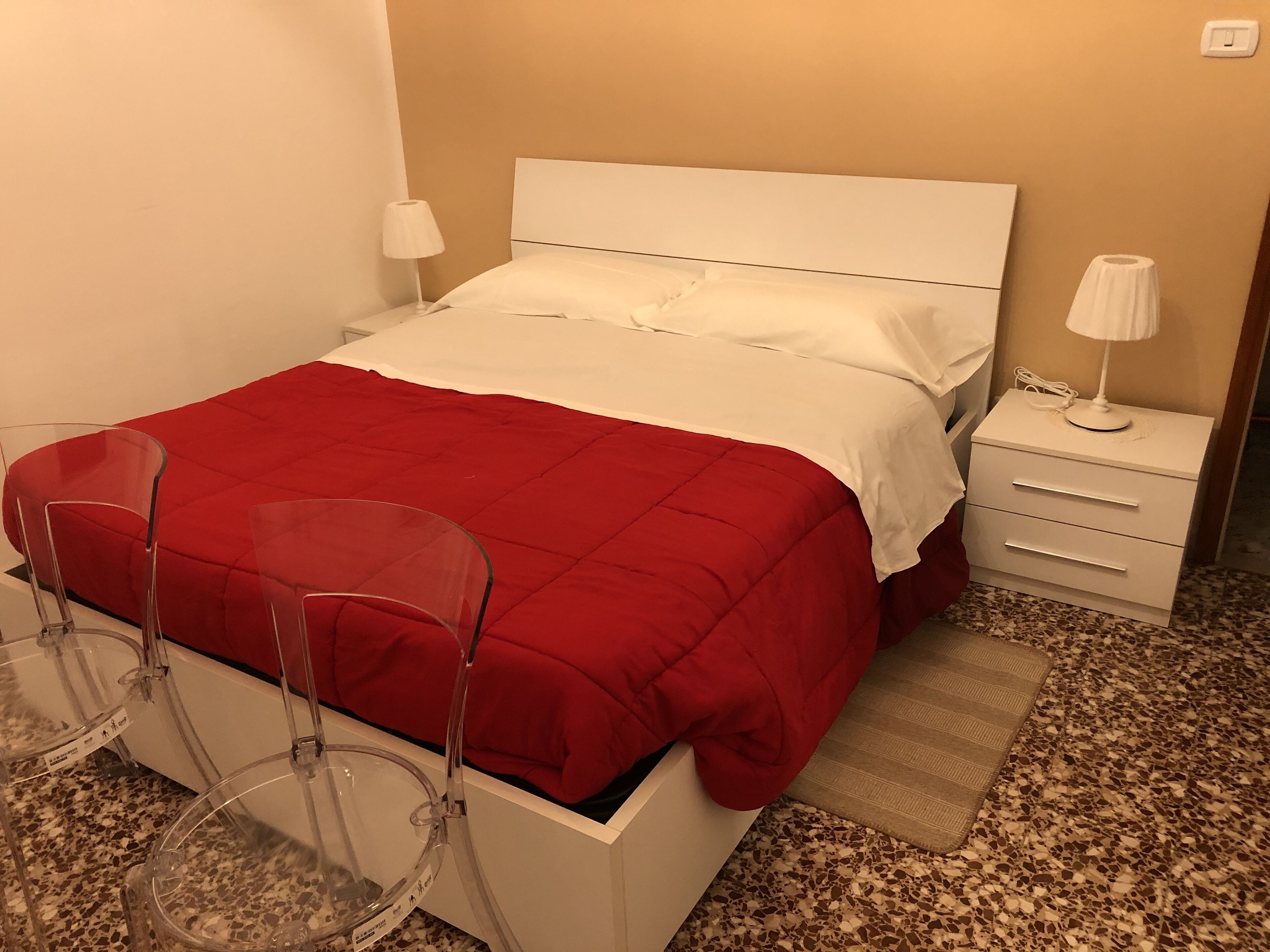 A neatly made bed with a red blanket, a white headboard, and a bedside table with a lamp. There are two clear chairs and a small rug on the floor.
