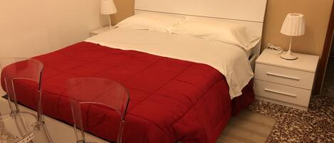 A neatly made bed with a red blanket, a white headboard, and a bedside table with a lamp. There are two clear chairs and a small rug on the floor.