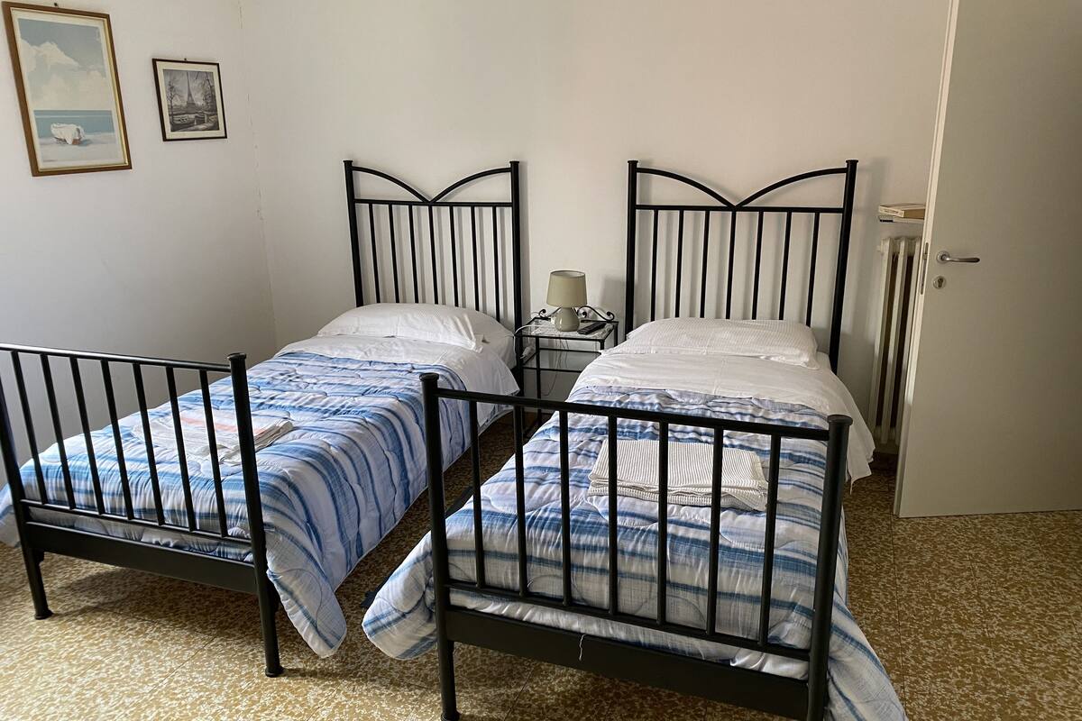 A room with two metal-framed beds with blue and white bedding, a bedside table with a lamp, and a door.