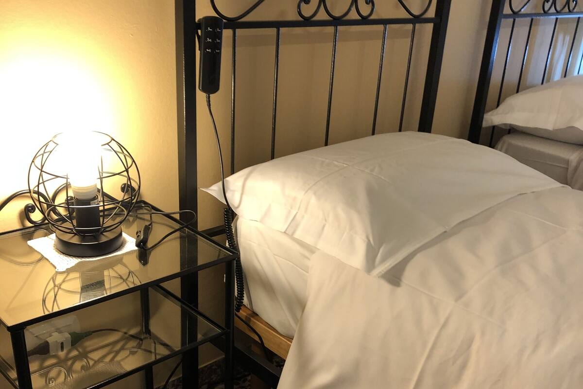 A single bed with white bedding, a metal headboard, and a glass table with a lamp.