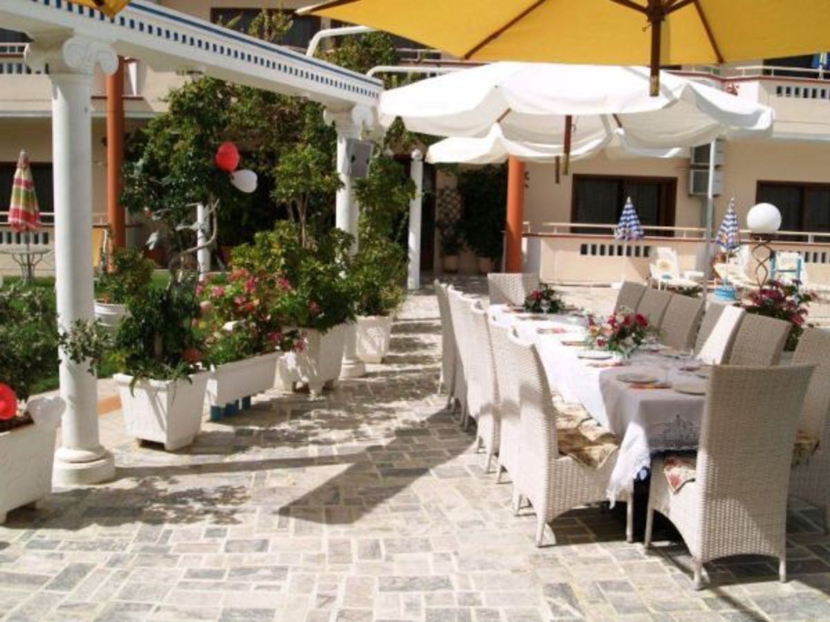 Un área de comedor al aire libre con mobiliario de mimbre blanco, plantas en macetas y un paraguas amarillo.