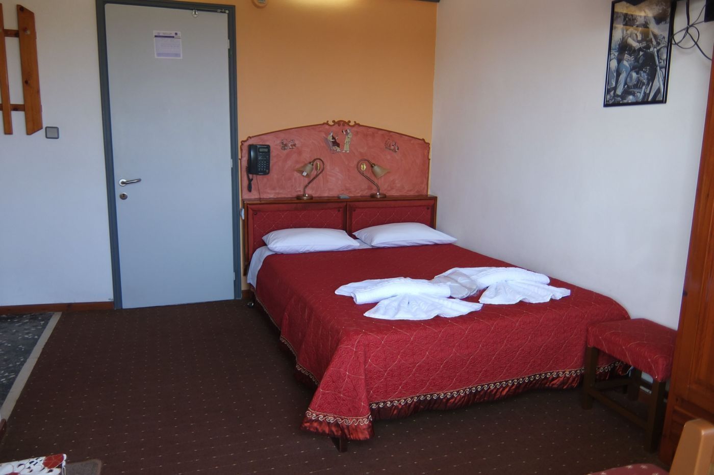 A hotel room with a single bed, a wooden door, a small desk, and a wall-mounted telephone.