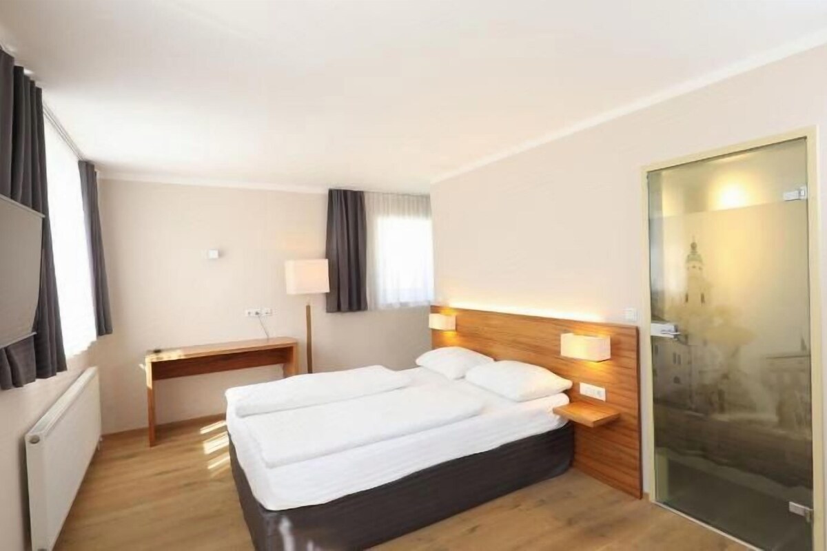A hotel room with a double bed, a desk, a chair, a mirror, and a view of a building through a glass door.