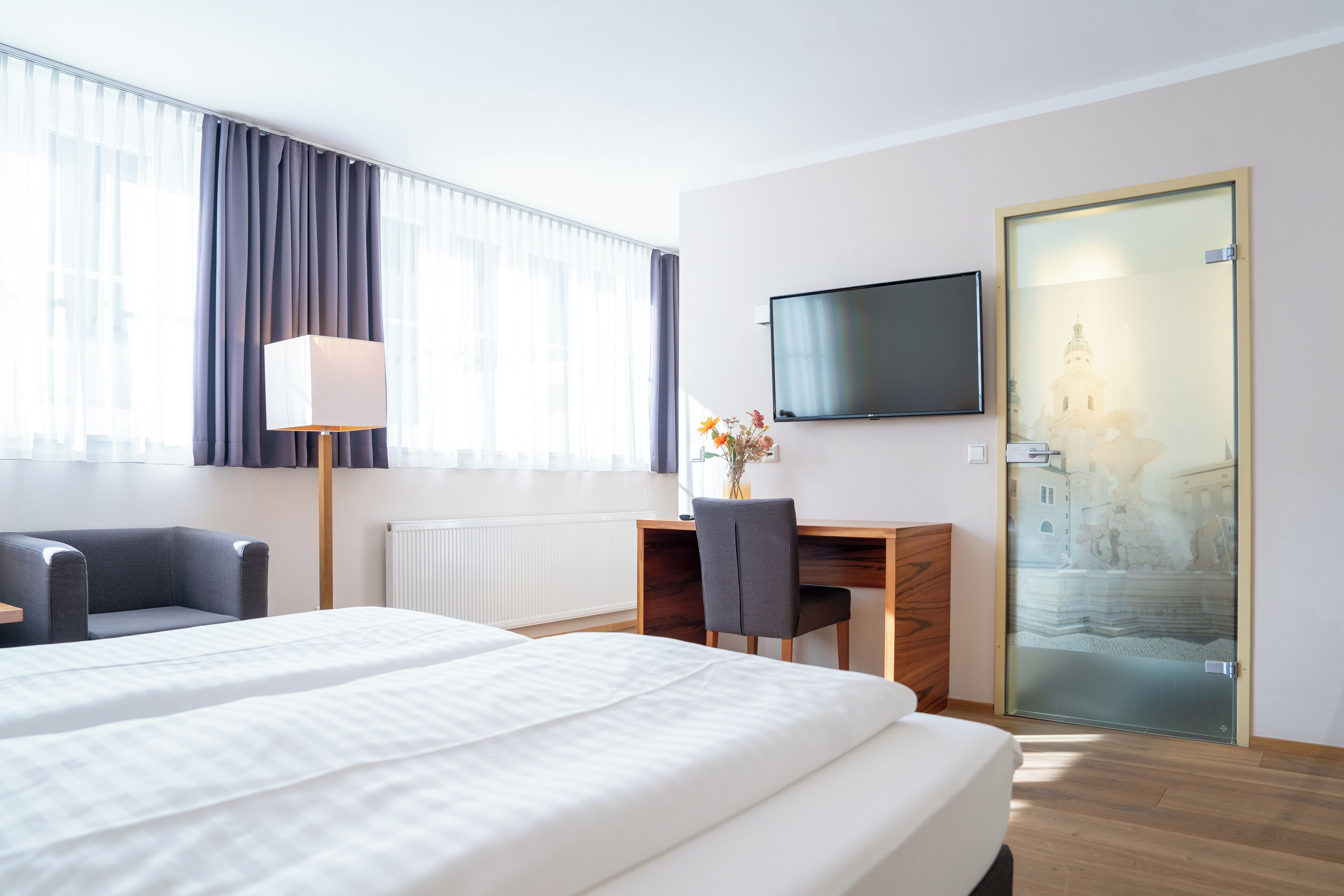 A hotel room with two beds, a desk, a chair, a TV, and a large window with curtains.
