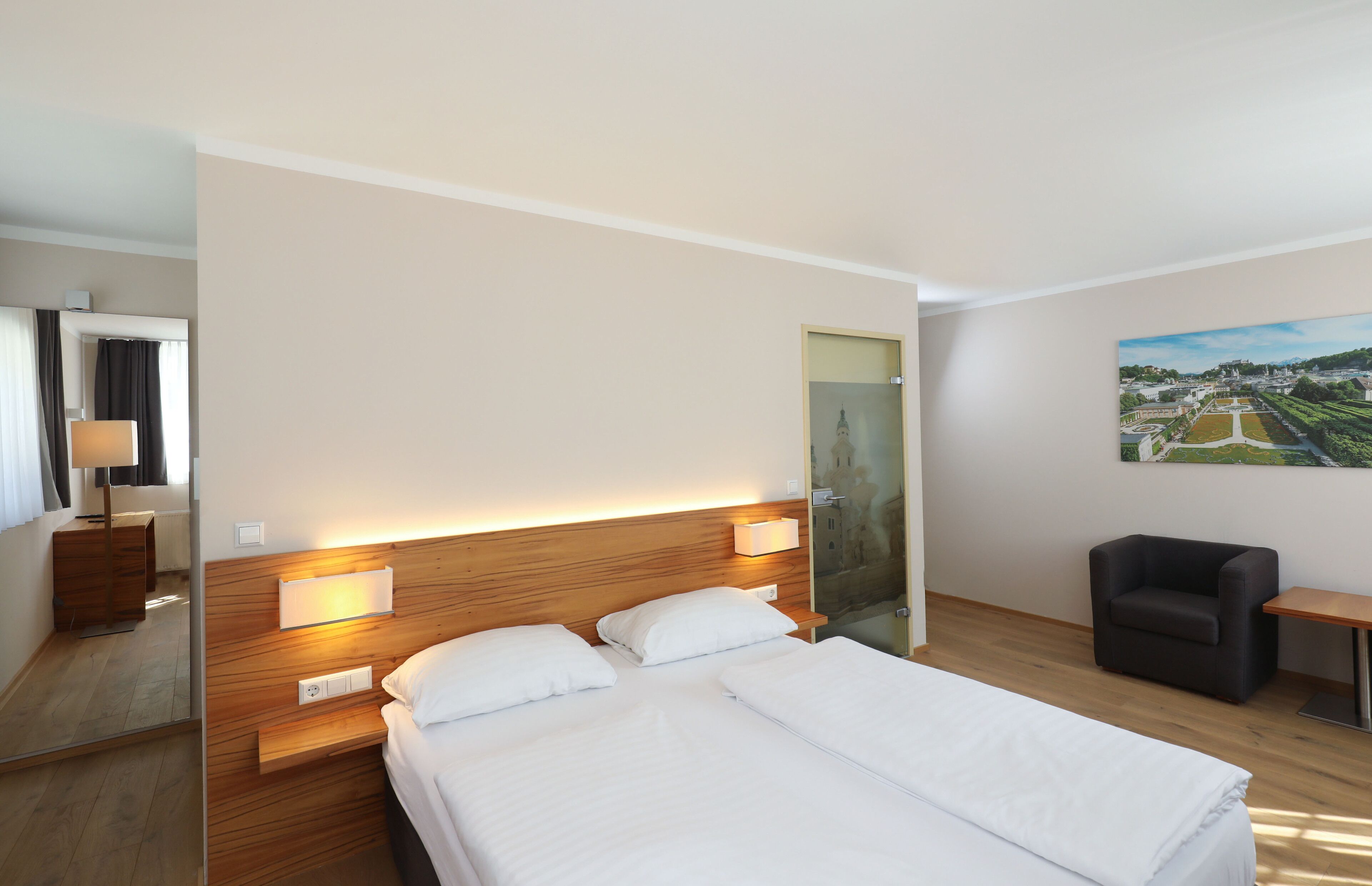 A hotel room with a large bed, bedside lamps, a small table, a chair, and a painting on the wall.