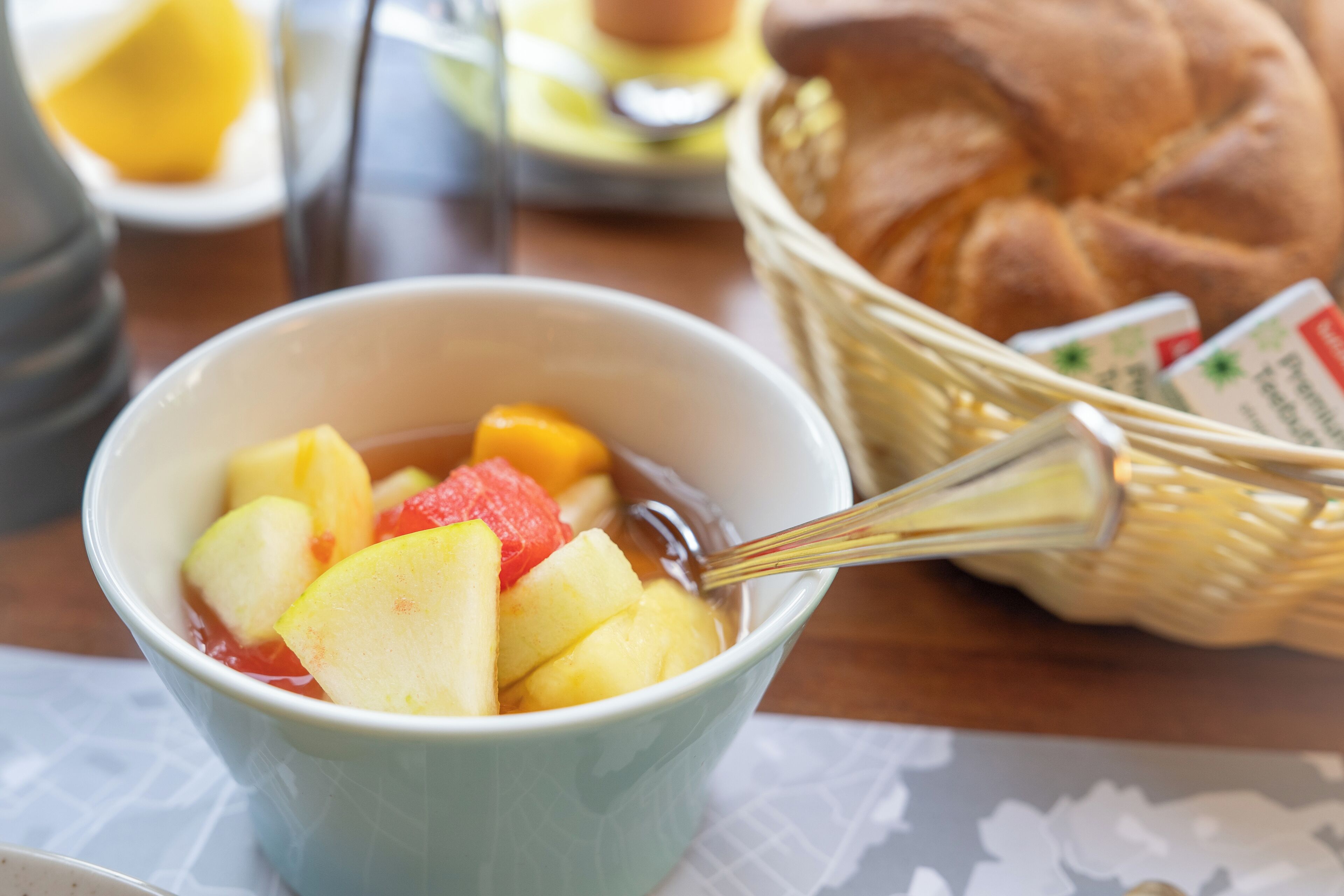 A bowl of fruit salad with a spoon, a basket of bread, and a cup of coffee on a table.
