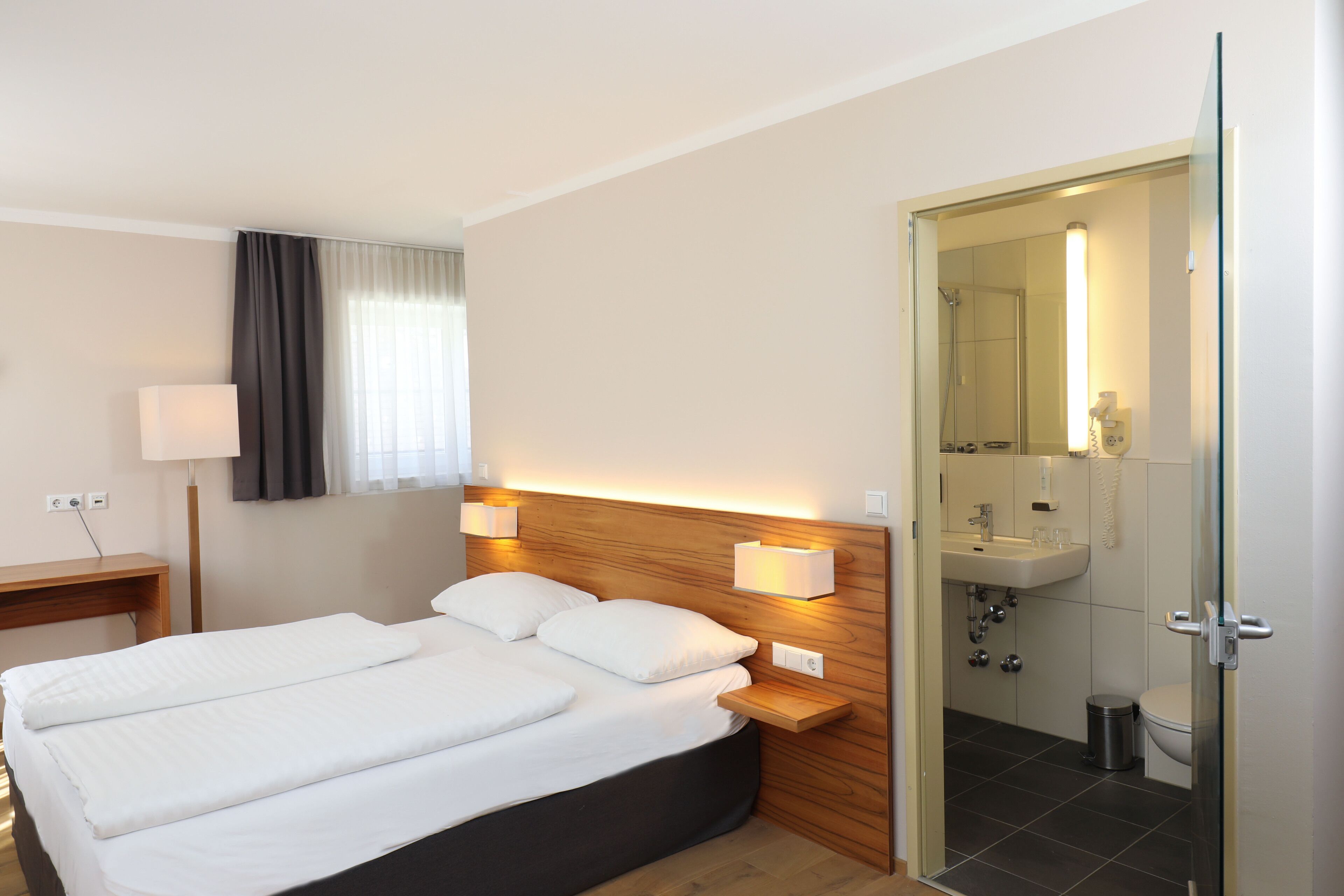A hotel room with a double bed, bedside lamps, a wooden headboard, a bedside table, a desk, a lamp, a window with curtains, and an open door leading to a bathroom with a sink, mirror, and toilet.
