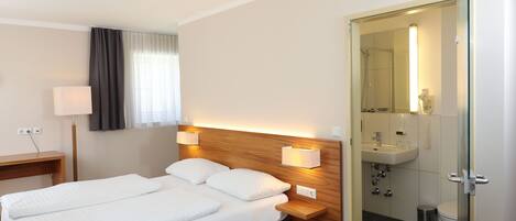 A hotel room with a double bed, bedside lamps, a wooden headboard, a bedside table, a desk, a lamp, a window with curtains, and an open door leading to a bathroom with a sink, mirror, and toilet.