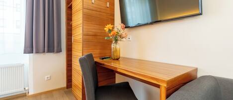 A room with a wooden desk, a grey chair, a TV mounted on the wall, and a window with curtains.