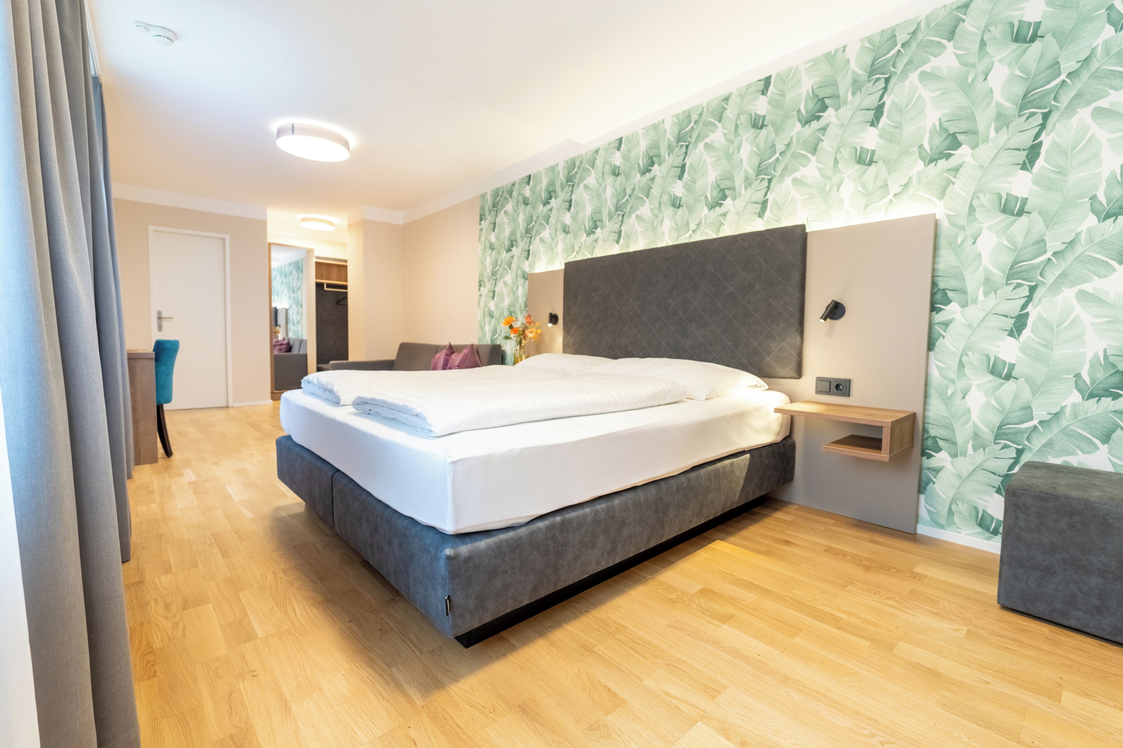 A modern hotel room with a large bed, a wooden floor, a grey sofa, and a wall with a leafy pattern.