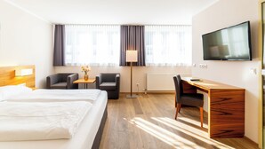 A hotel room with two beds, a desk, a chair, a TV, and large windows with curtains. - Das Jedermann Boutiquehotel (Salzburg)