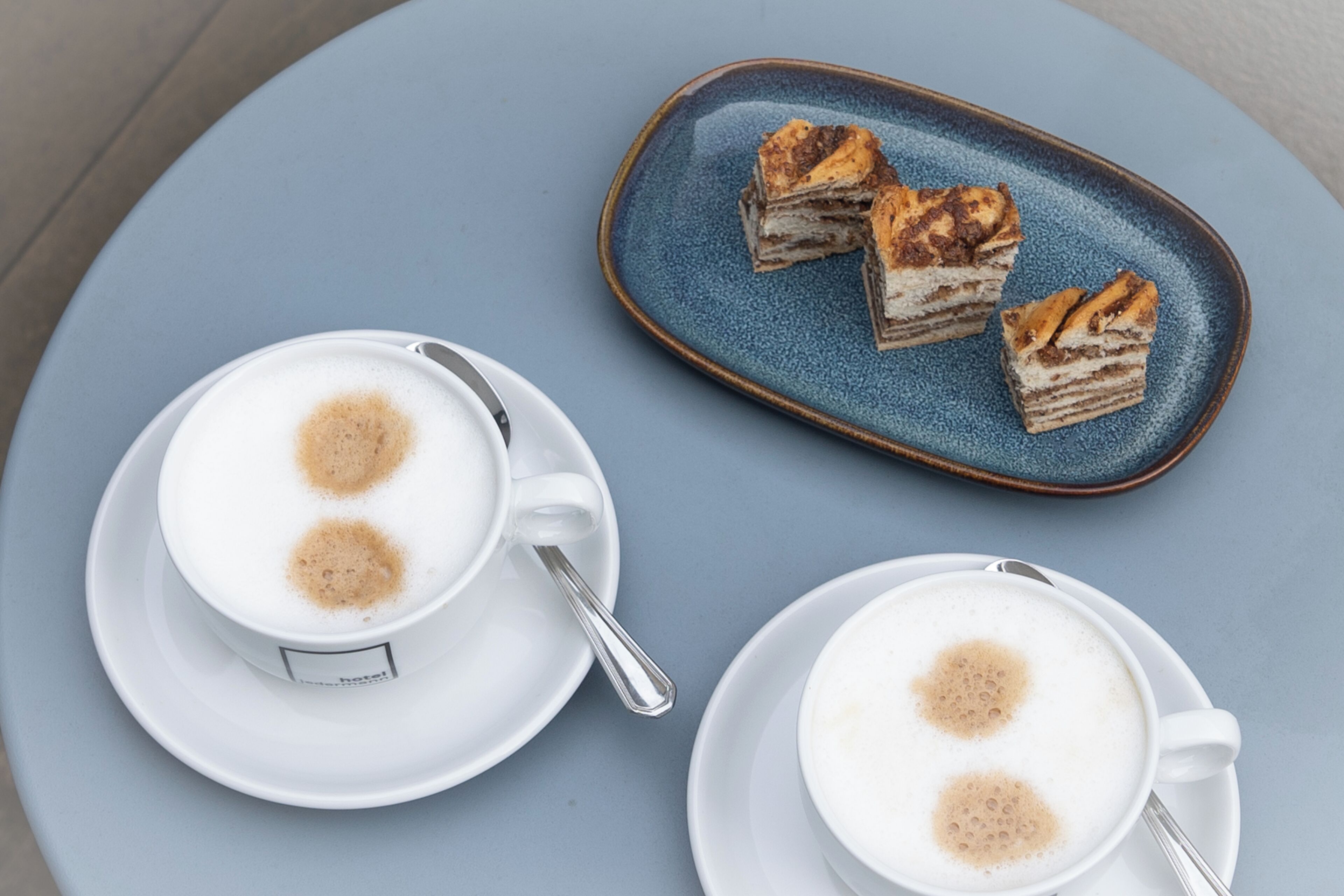 Two cups of coffee with foam on a blue table, and a plate with slices of cake.