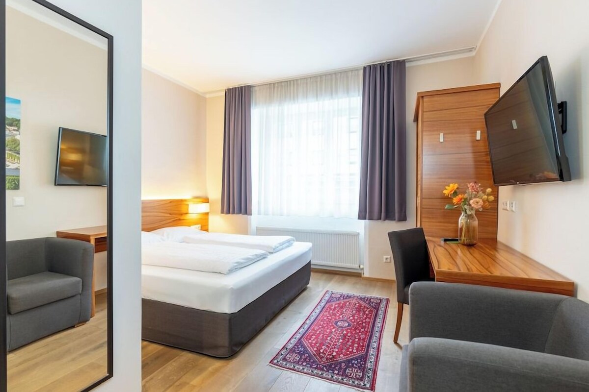 A hotel room with a bed, a desk, a chair, a TV, and a window with curtains.