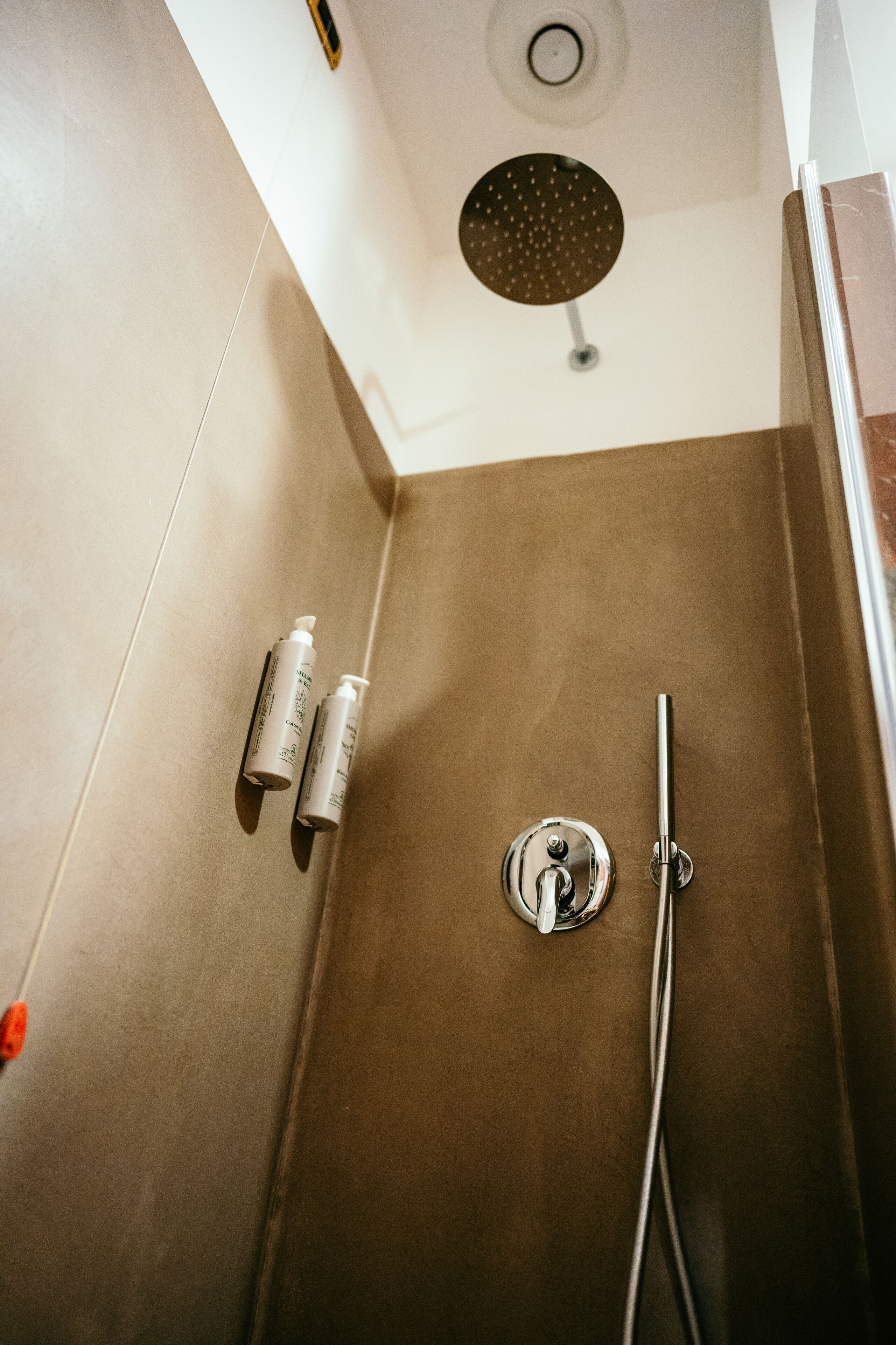 A shower area with a brown wall, a ceiling-mounted showerhead, and two bottles of shampoo on the wall.