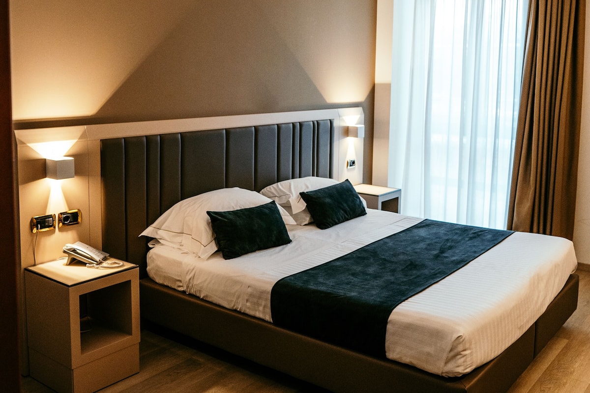Une chambre d’hôtel moderne avec un grand lit, une table de chevet avec une lampe et une vue sur la ville par la fenêtre.