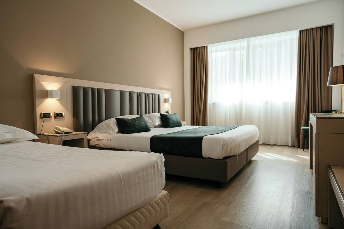 A hotel room with two beds, a desk with a lamp, a phone, and a large window with curtains.