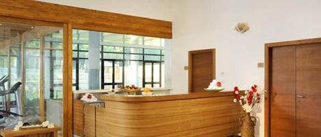 A reception area with a wooden reception desk, a bench, a vase with flowers, and a view through large windows.