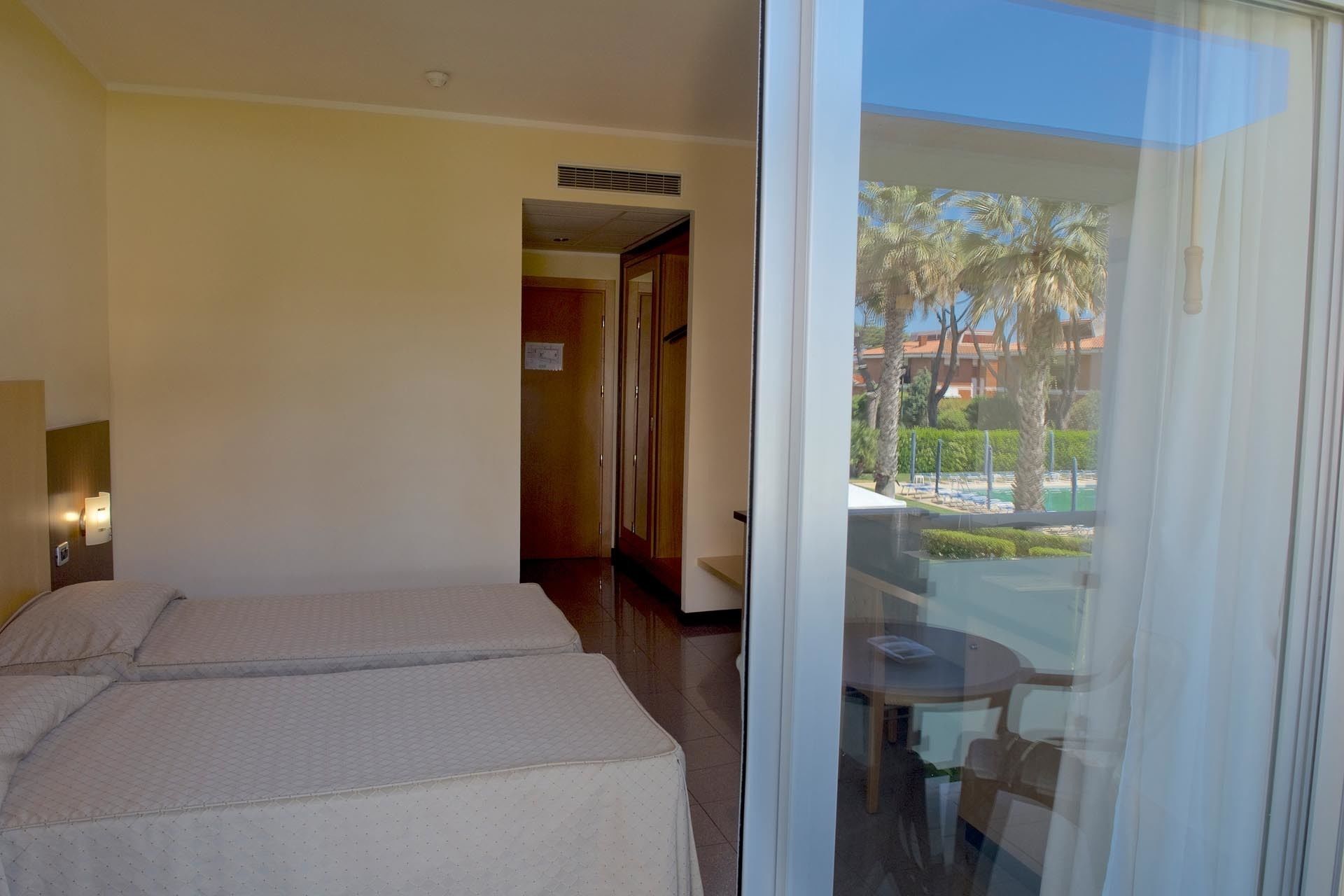 A hotel room with two beds, a small table, and a view of a pool and palm trees through a glass door.