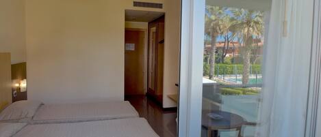 A hotel room with two beds, a small table, and a view of a pool and palm trees through a glass door.
