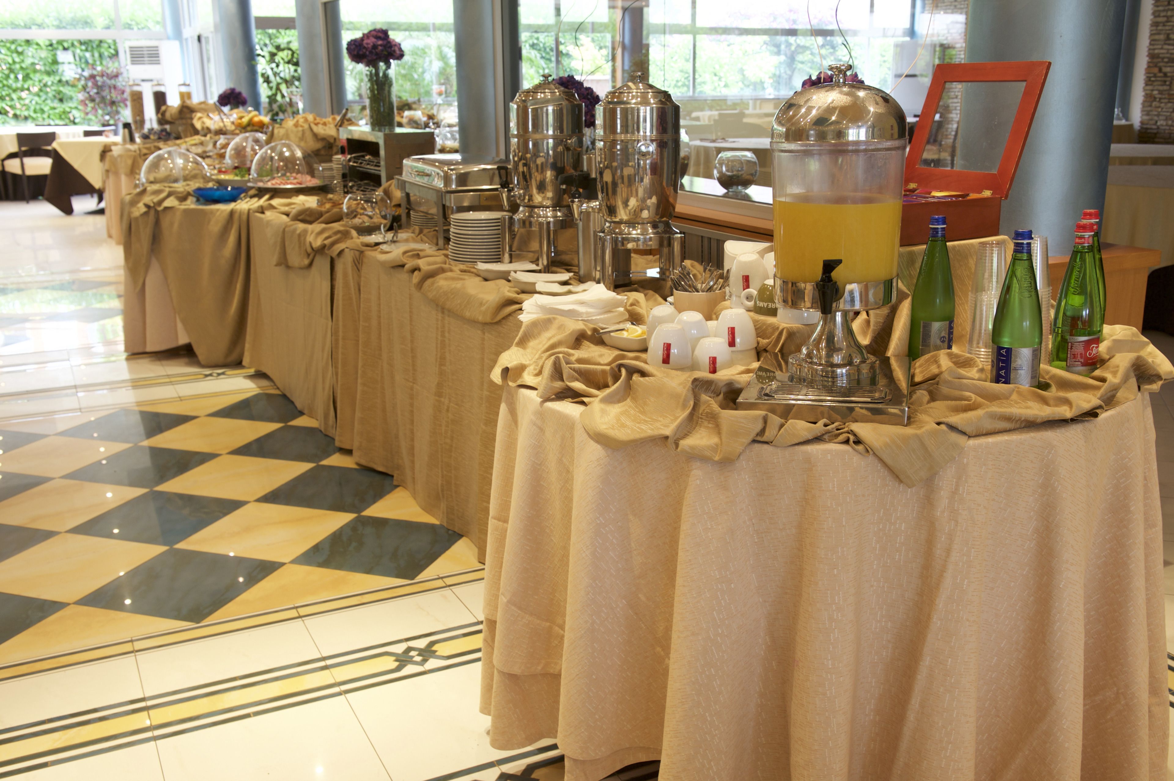 A buffet setup with multiple tables covered in beige cloths, featuring various food and drink dispensers, plates, and glassware.