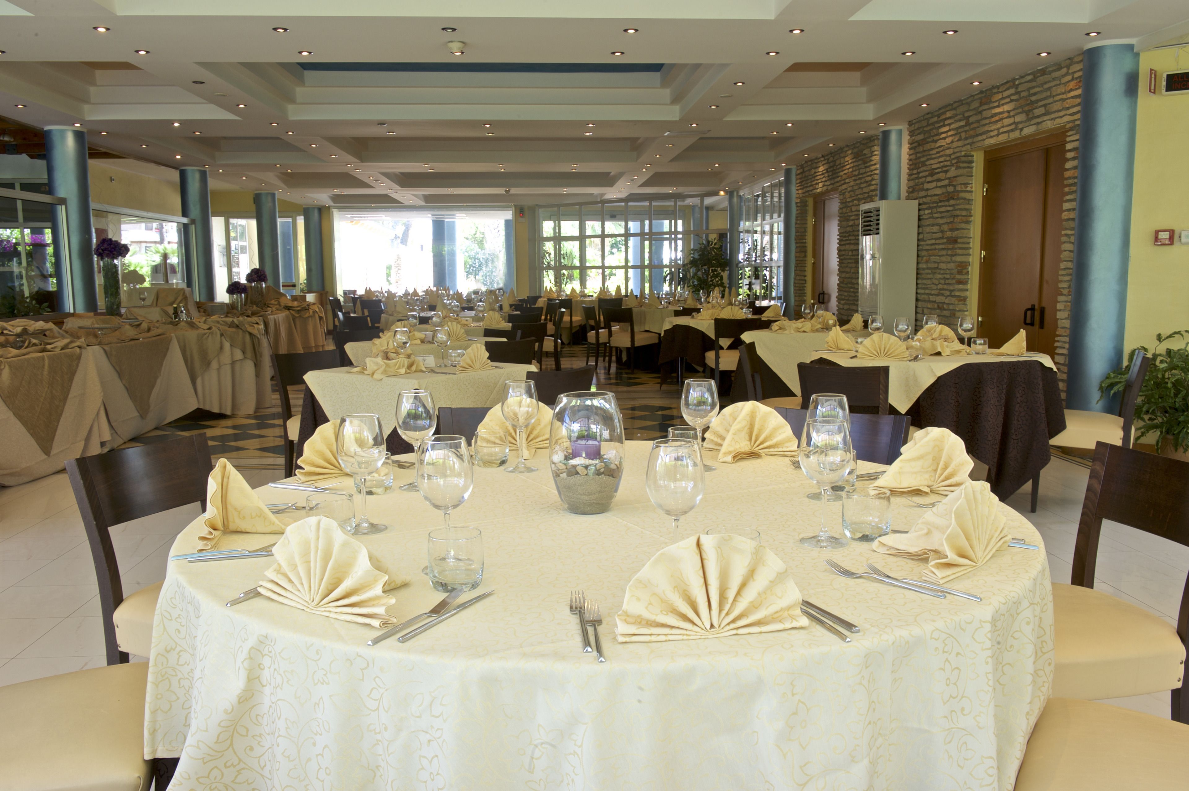 Un vaste espace dédié à la salle à manger, avec des tables rondes dressées pour le repas, recouvertes de nappes blanches et agrémentées d’une vaisselle soigneusement disposée.