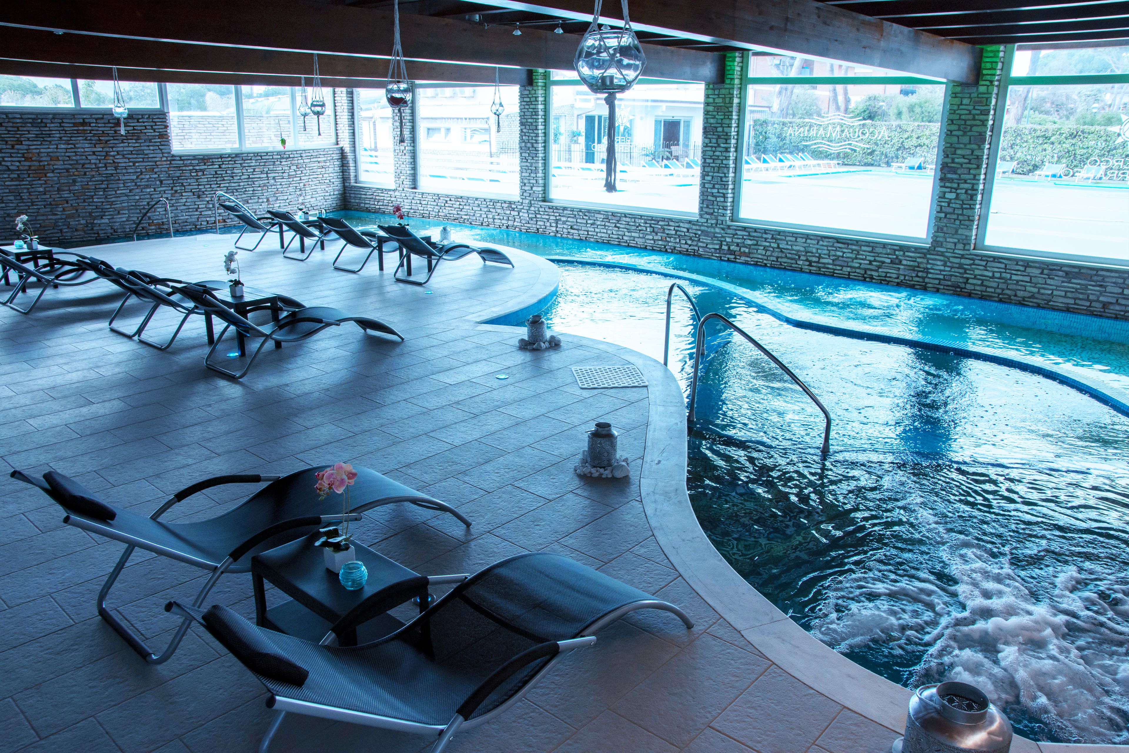 Una piscina interior con sillas de descanso y una mesa, ventanas amplias y una pared de ladrillo.