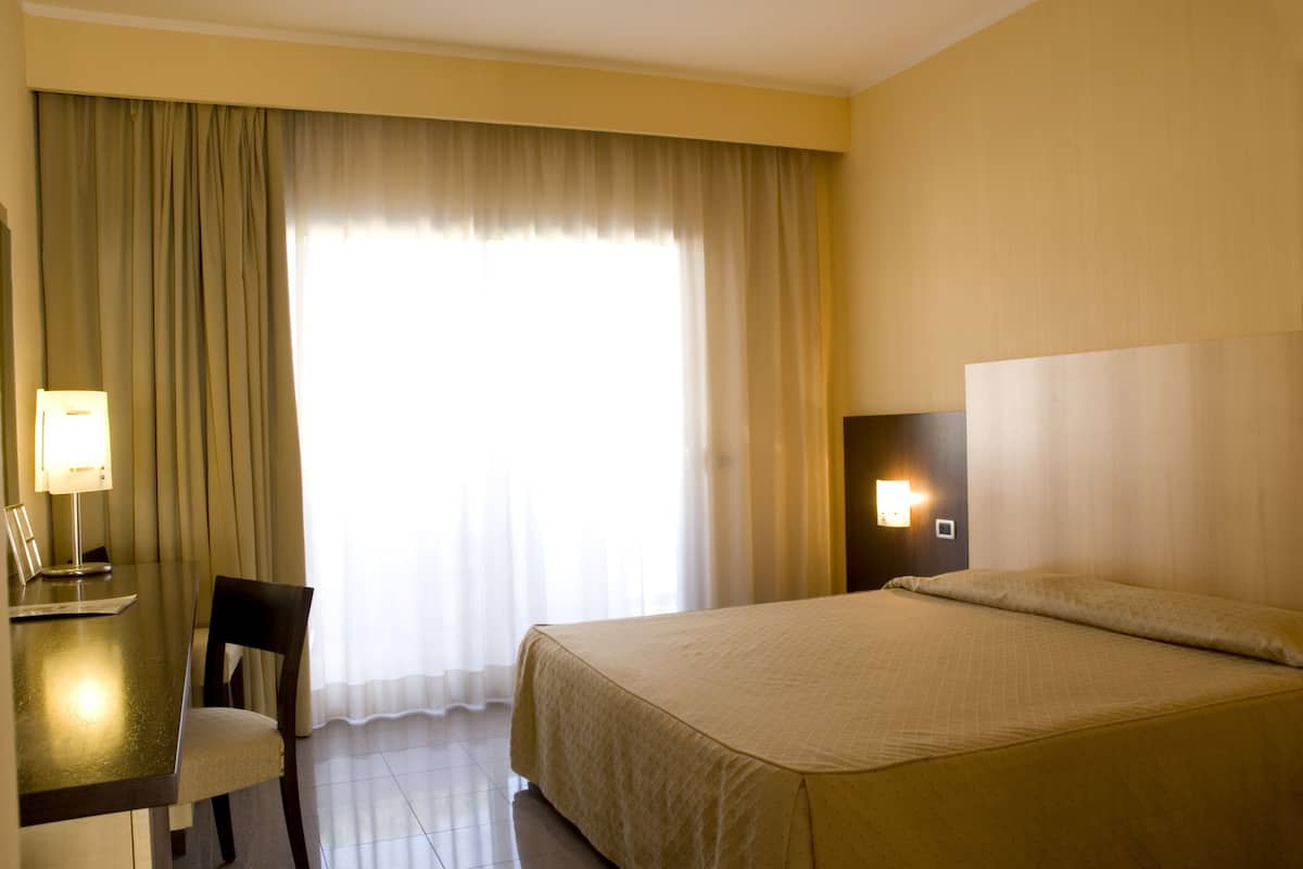 A hotel room with a bed, a desk with a chair, a lamp, and a window with curtains.