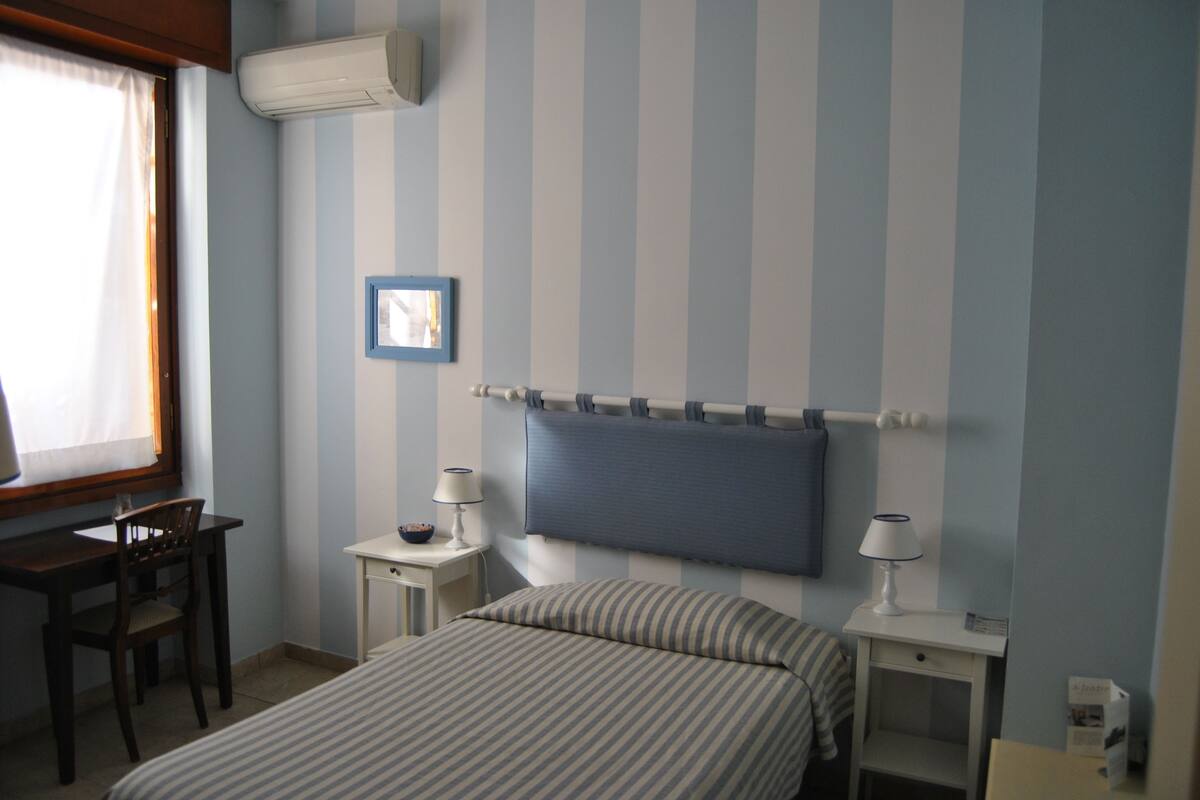 A hotel room with a bed, two bedside tables with lamps, a desk with a chair, and a window with curtains.