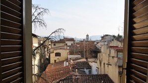 A view from a balcony with wooden shutters, overlooking a densely packed urban area with terracotta-tiled roofs and traditional architecture.