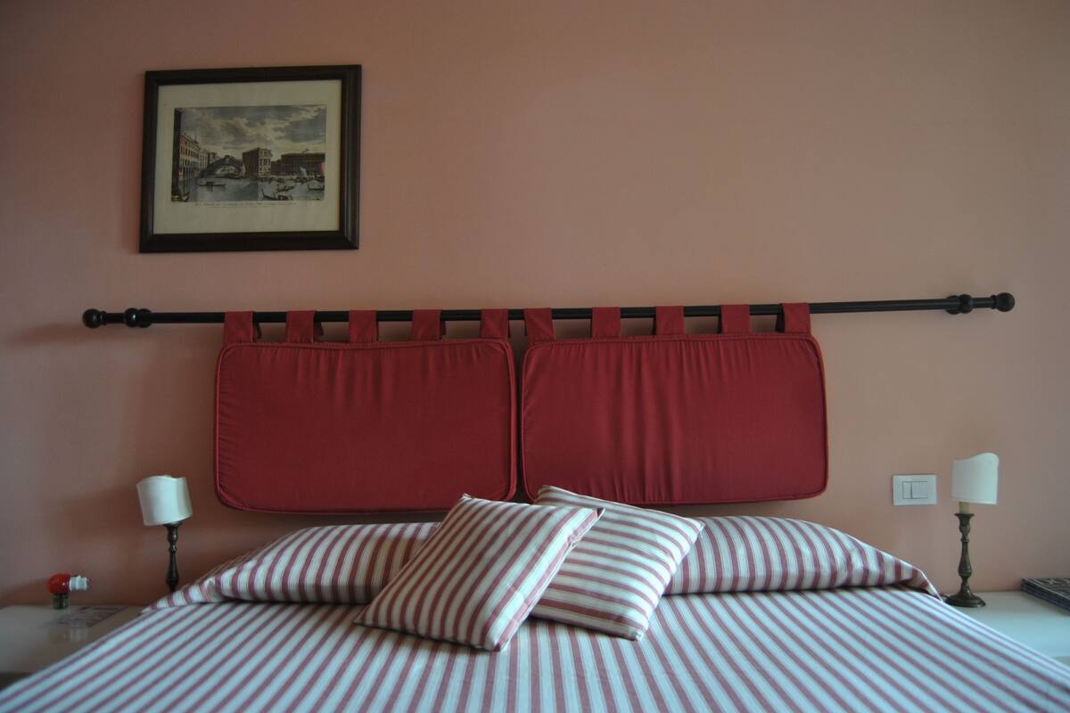 A neatly made bed with striped bedding, a headboard with red curtains, a framed picture on the wall, and bedside lamps.