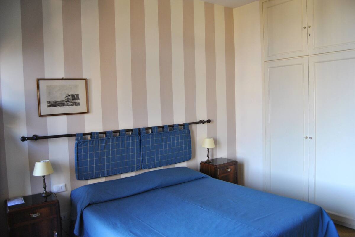A bedroom with a striped wall, a bed with blue bedding, a nightstand with a lamp, and a wardrobe.
