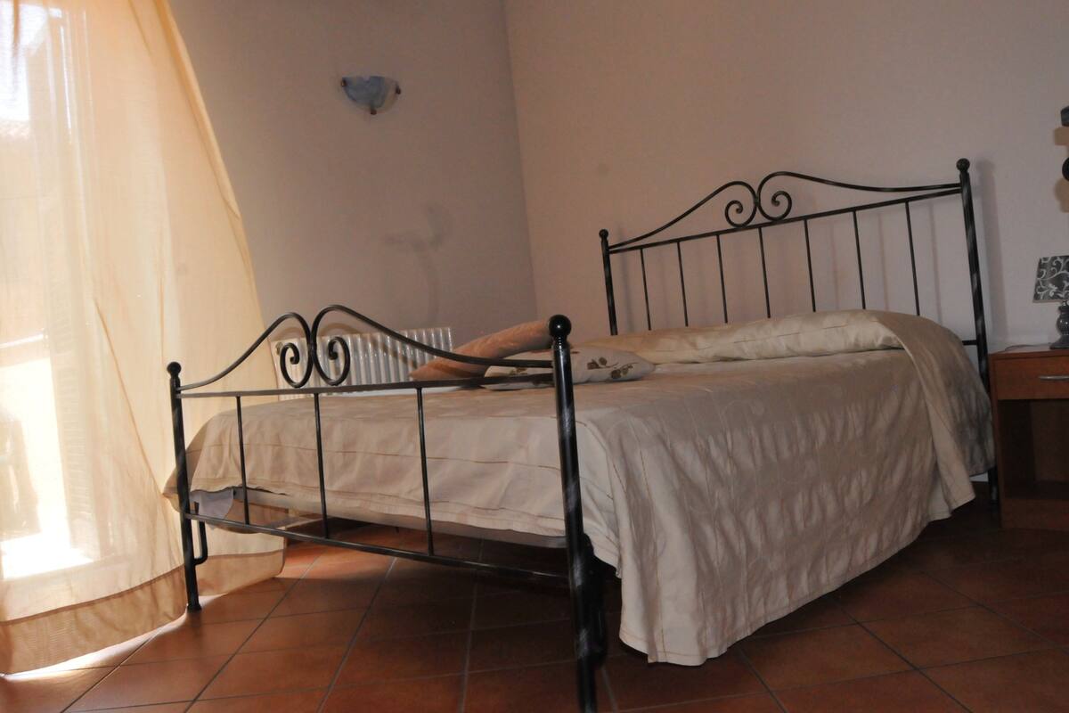 Ein Einzelbett mit Metallgestell, einer beigen Tagesdecke und einem Nachttisch mit Lampe.