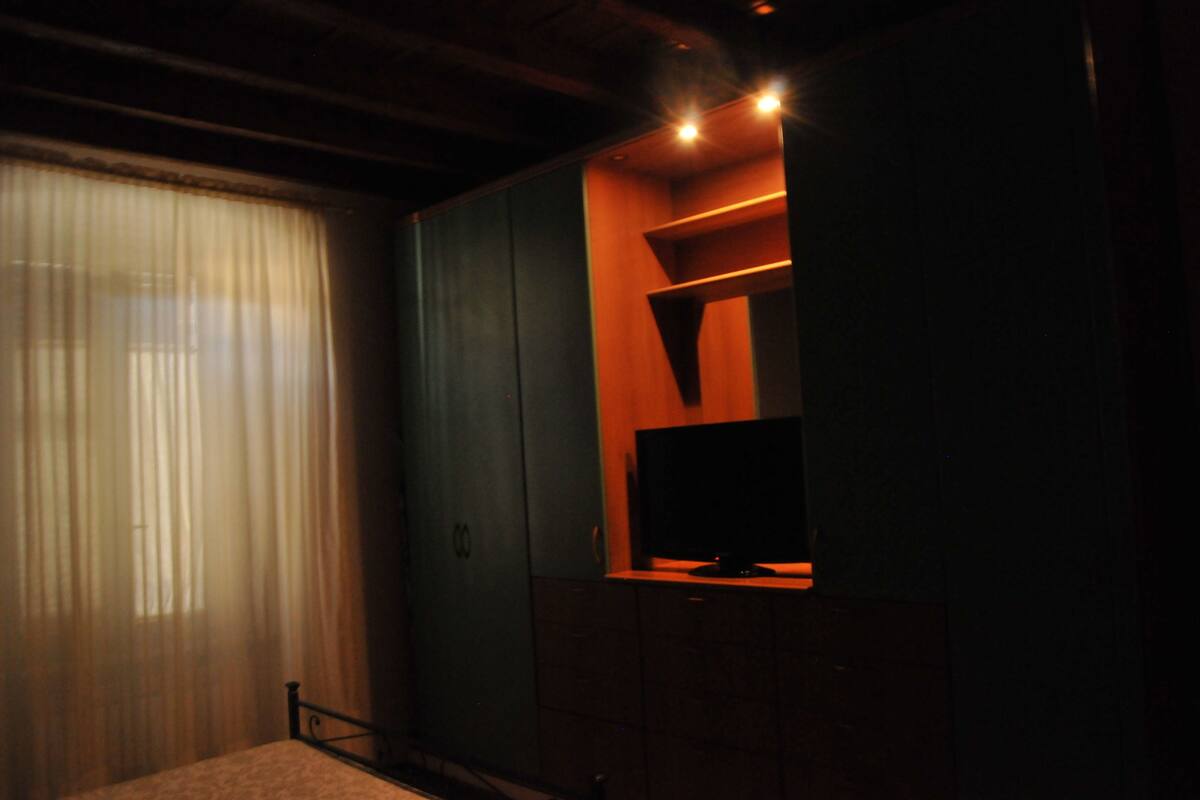 Ein schwach beleuchteter Raum mit einem großen Fenster, das mit transparenten Vorhängen bedeckt ist, einem Fernseher auf einem Holzständer und einem kleinen Bett mit Metallgestell.
