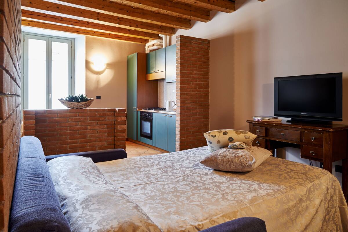 Ein gemütliches Zimmer mit Bett, Fernseher und einer Küchenzeile mit Backsteinwand und Holzbalken.