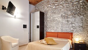 Um quarto com parede de pedra, cama com cabeceira laranja, poltrona branca e televisão fixada na parede.