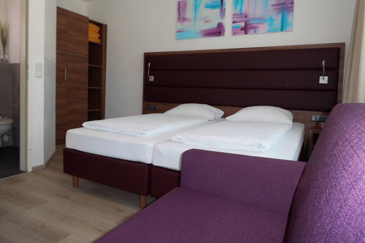Hotellihuone, jossa on sänky, violetti sohva, puinen vaatekaappi ja abstrakti seinätaide.