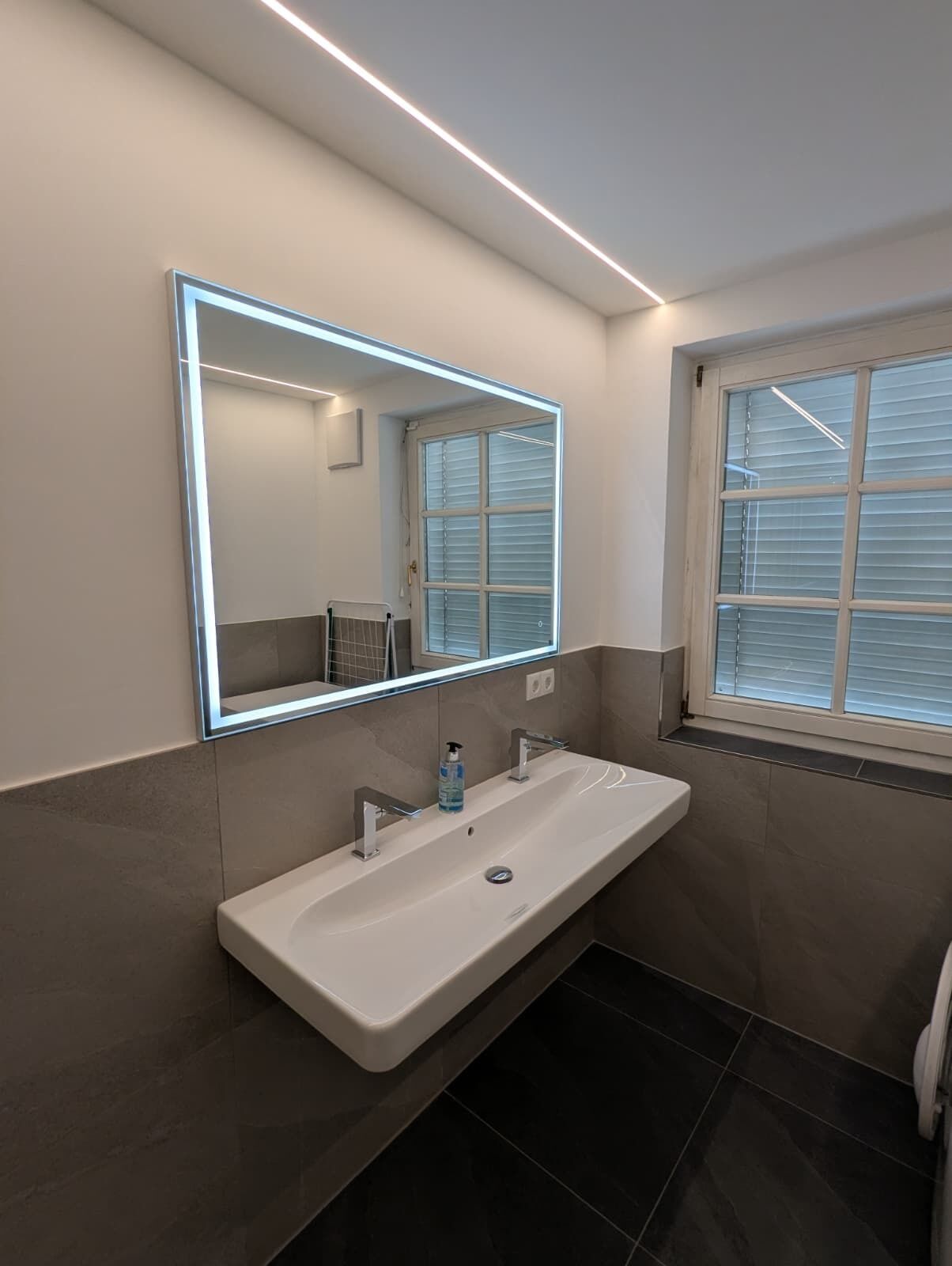 Une salle de bain moderne avec un grand miroir, un lavabo blanc et une fenêtre équipée de stores.