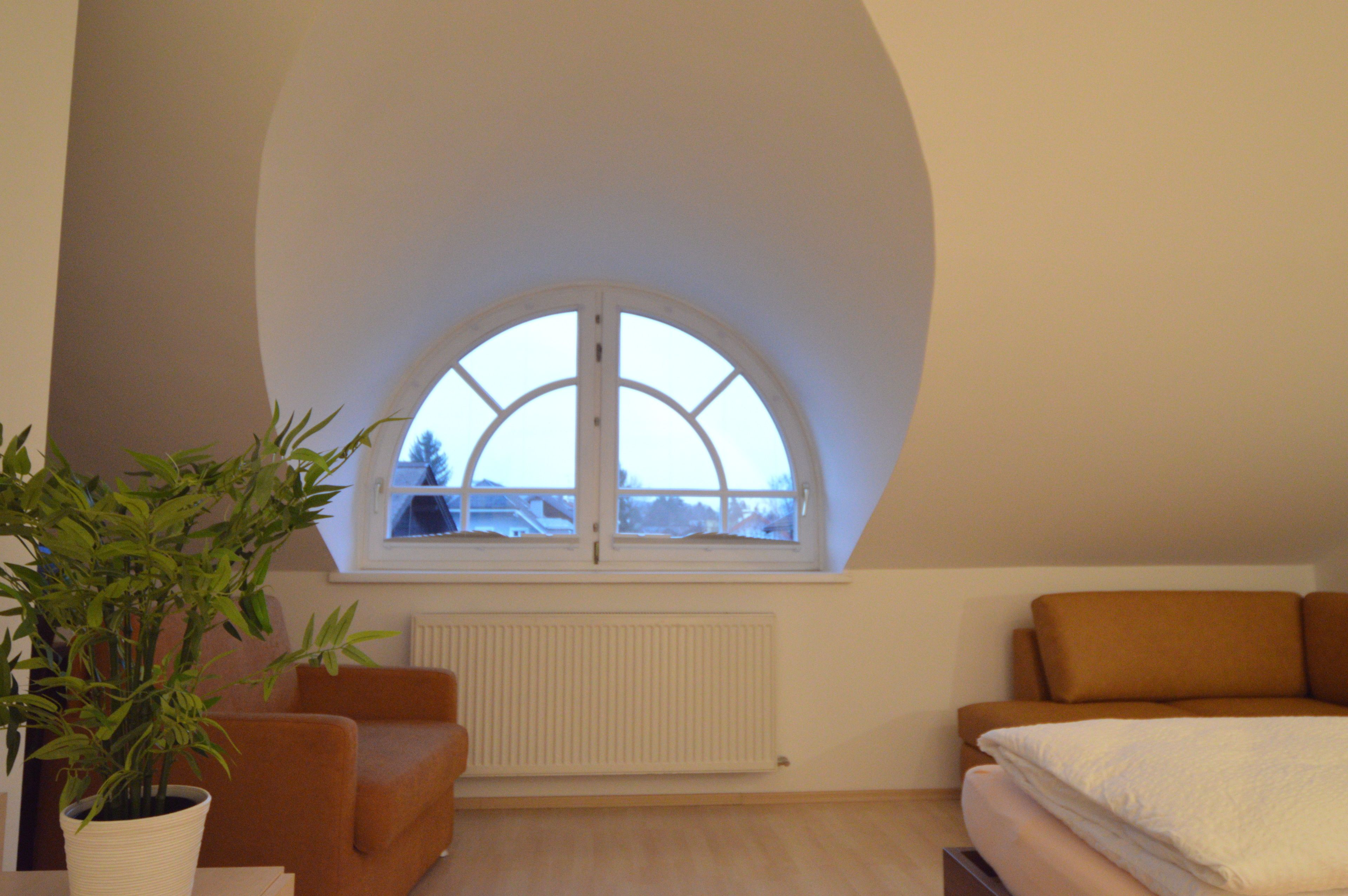 A room with a curved ceiling, a large arched window, a radiator, a brown sofa, a white bed, and a potted plant.